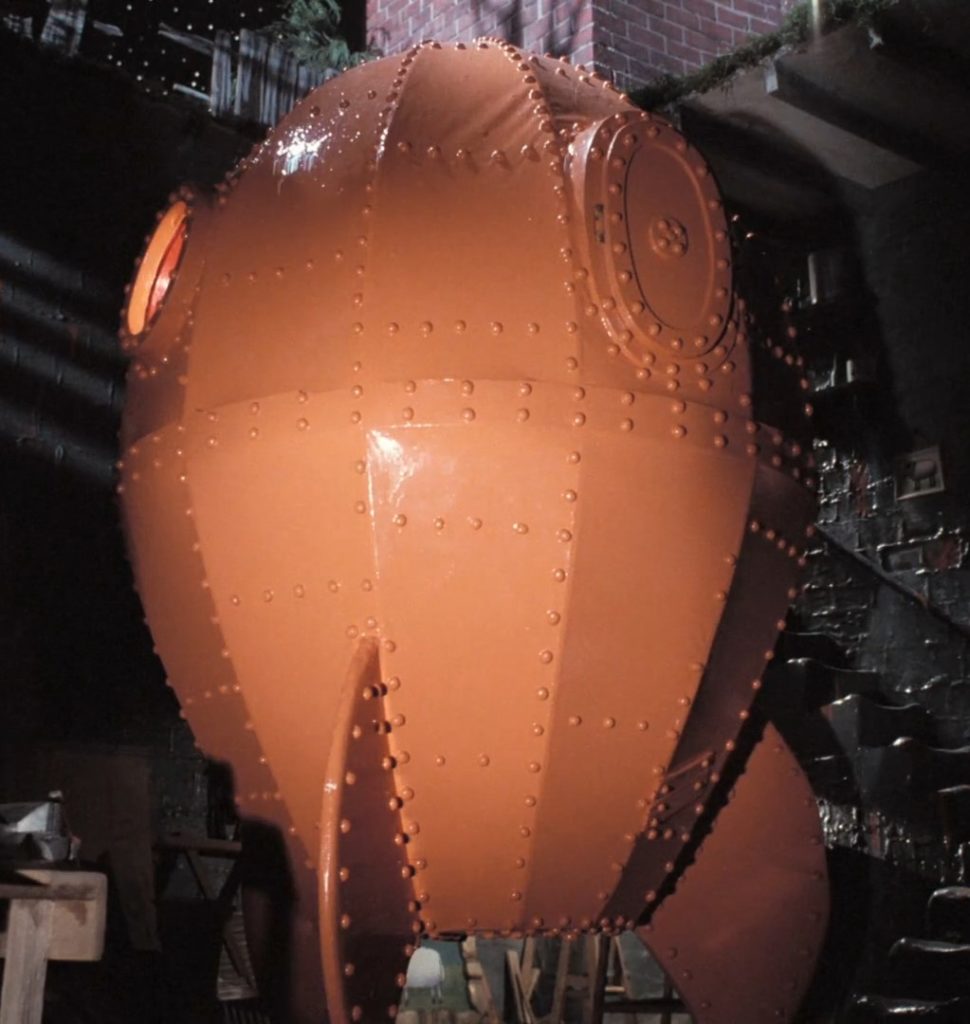 A bulbous orange rocketship with fins and prominent rivets in a basement. There is a single porthole visible and a hatch that would require a ladder to reach.