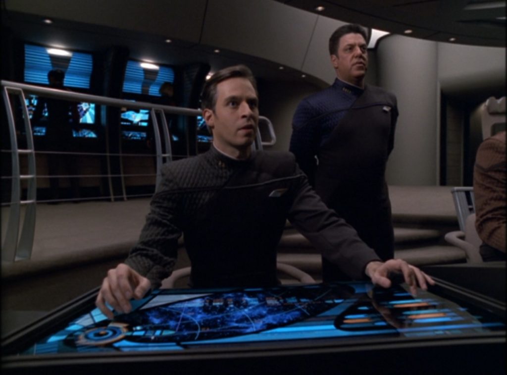 Characters in a science-fiction room with gray carpet and walls, metal railings, and blue computer interfaces.