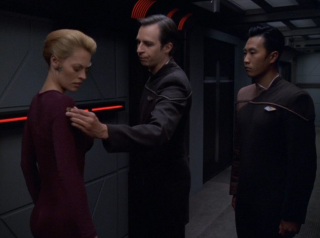 Two men in futuristic uniforms place a small device on Seven of Nine's upper arm.