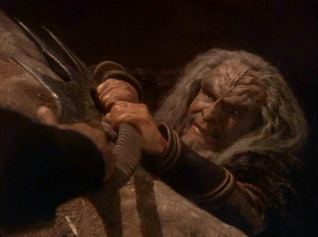 A gray-haired Klingon grips a curved bat'leth with both hands. From off-camera, a uniformed hand holds the other end of the sword.