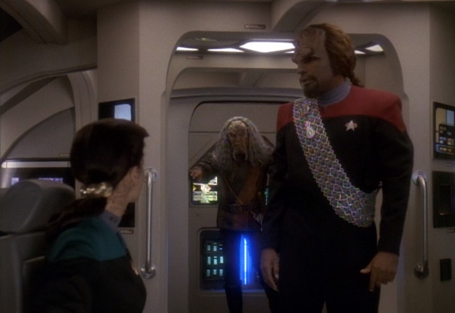 Jadzia Dax is in the foreground, with Worf in the middleground and Kor emerging from a doorway in the background.