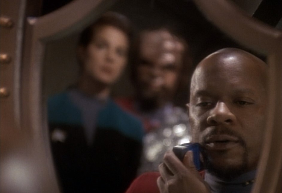 A Black man with a shaved head trims his beard in a mirror. Jadzia Dax and Worf are out of focus in the background of the reflection.