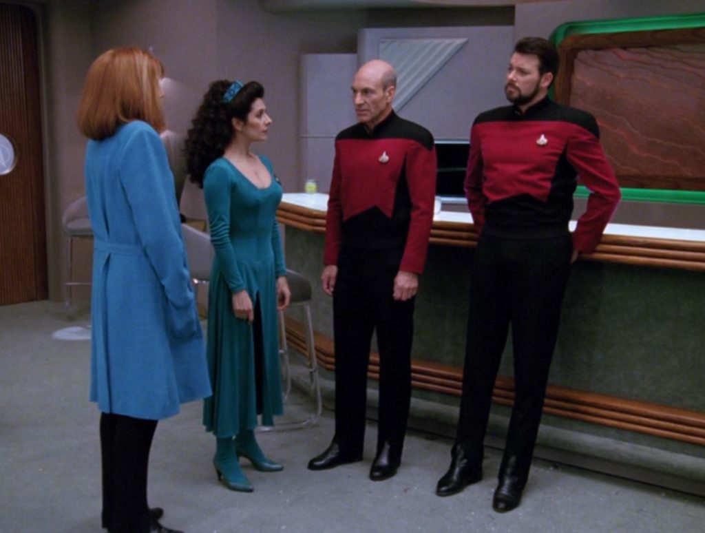 Counselor Troi, a dark-haired woman, is wearing a dark teal dress with long sleeves, an asymmetrical neckline and a slit skirt, as well as tights and high-heeled shoes in the same color. With her are Capt. Picard, Cmdr. Riker, and Dr. Crusher in standard uniforms. Crusher wears a soft blue doctor's coat over hers.