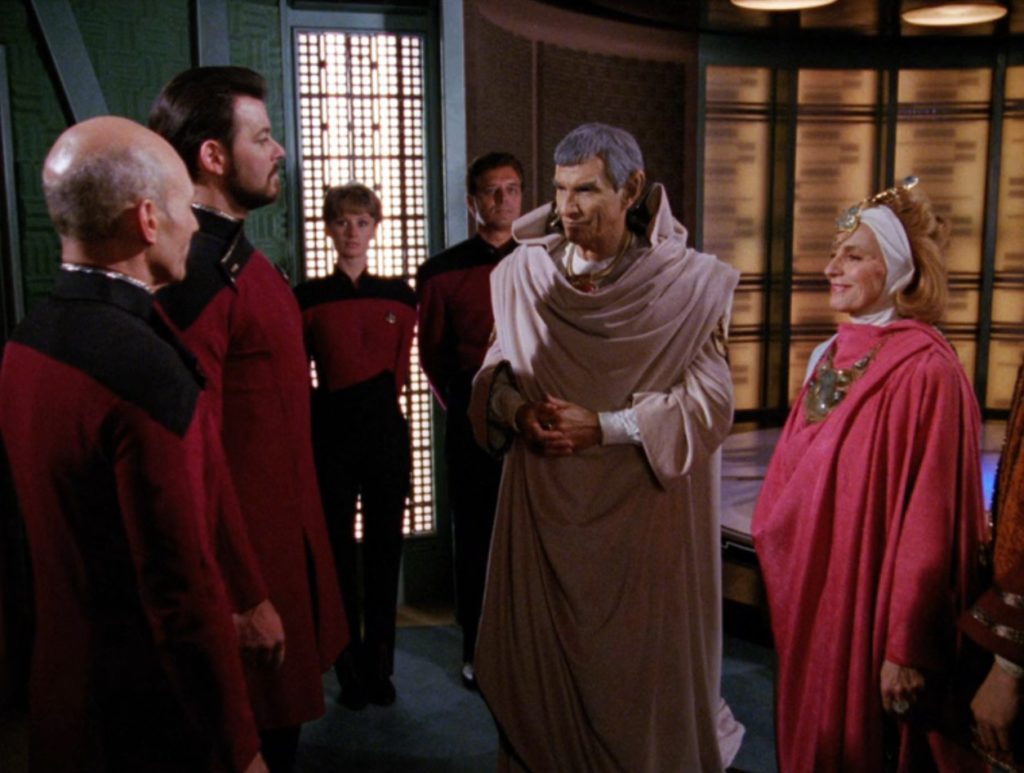 Picard and Riker (wearing thigh-length red tunics over black tights) greet Sarek (wearing voluminous taupe robes) and Perrin (wearing red robes, a white open-backed cowl, and prominent neck and forehead jewelry).