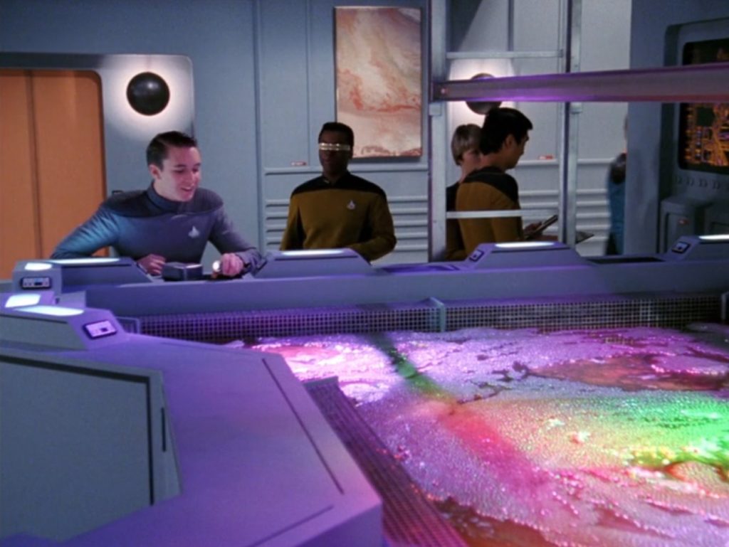 Wesley Crusher and Geordie LaForge examine a rainbow-lit bubbling pool in a dim meeting chamber.