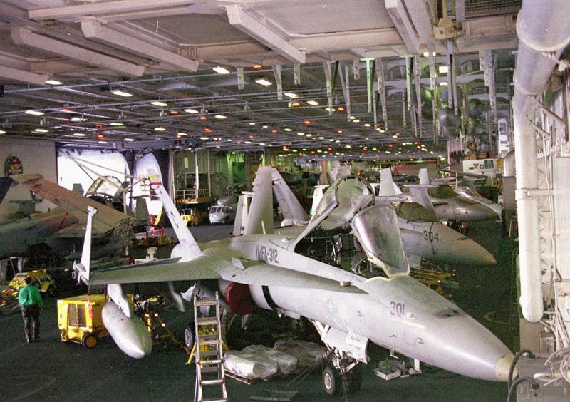 A hangar with a low ceiling supported by visible structural members, a gray industrial-looking floor, and planes lining the long walls.