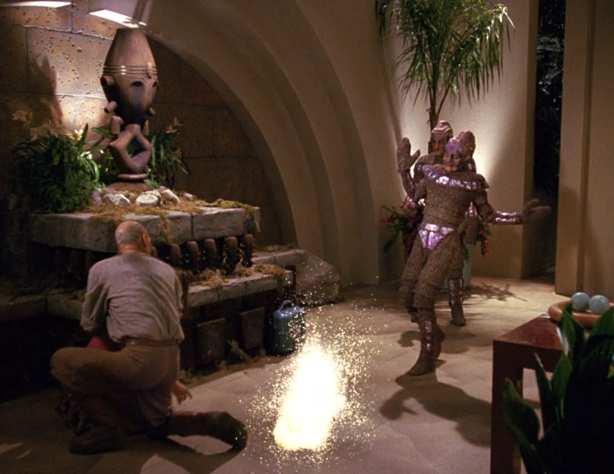 An alien in brown and shiny purple recoils from a small explosion as Picard shields Vash.